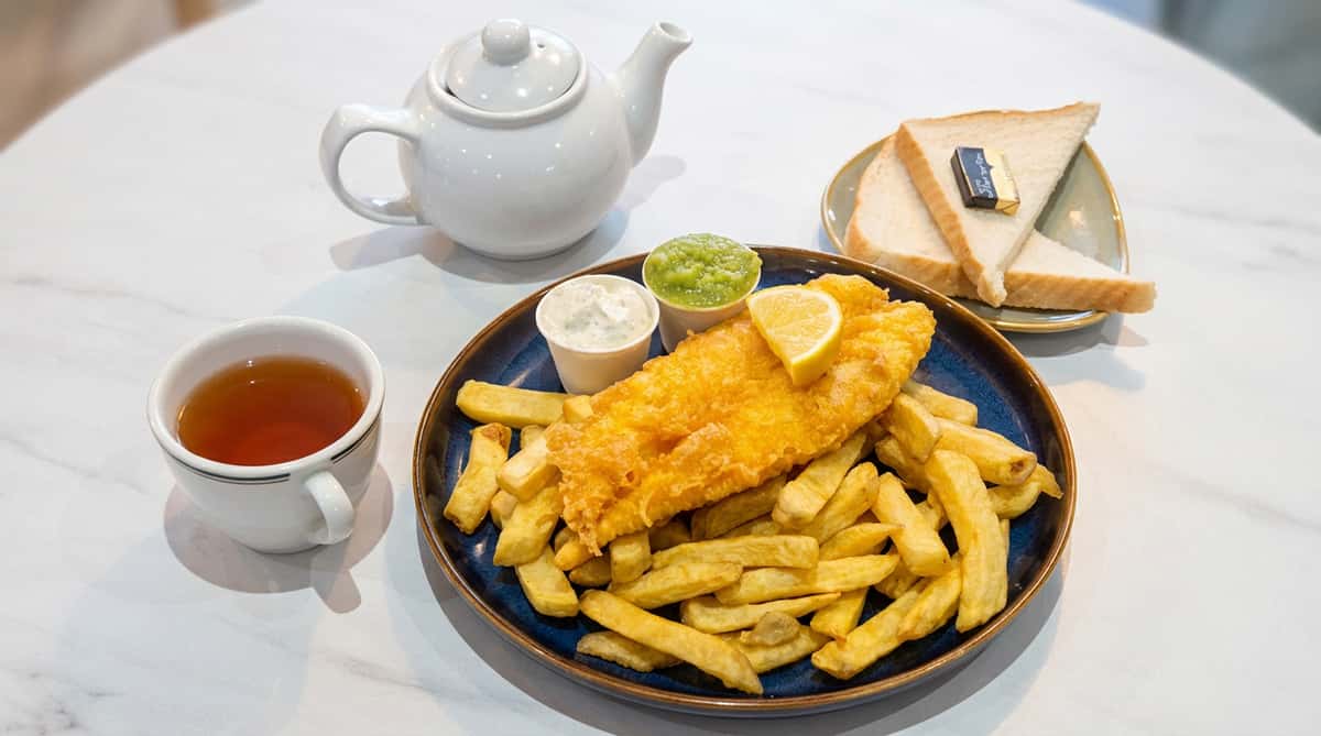 Fish and chips with a lemon wedge, tartar sauce, mushy peas, tea, and sliced bread with butter.