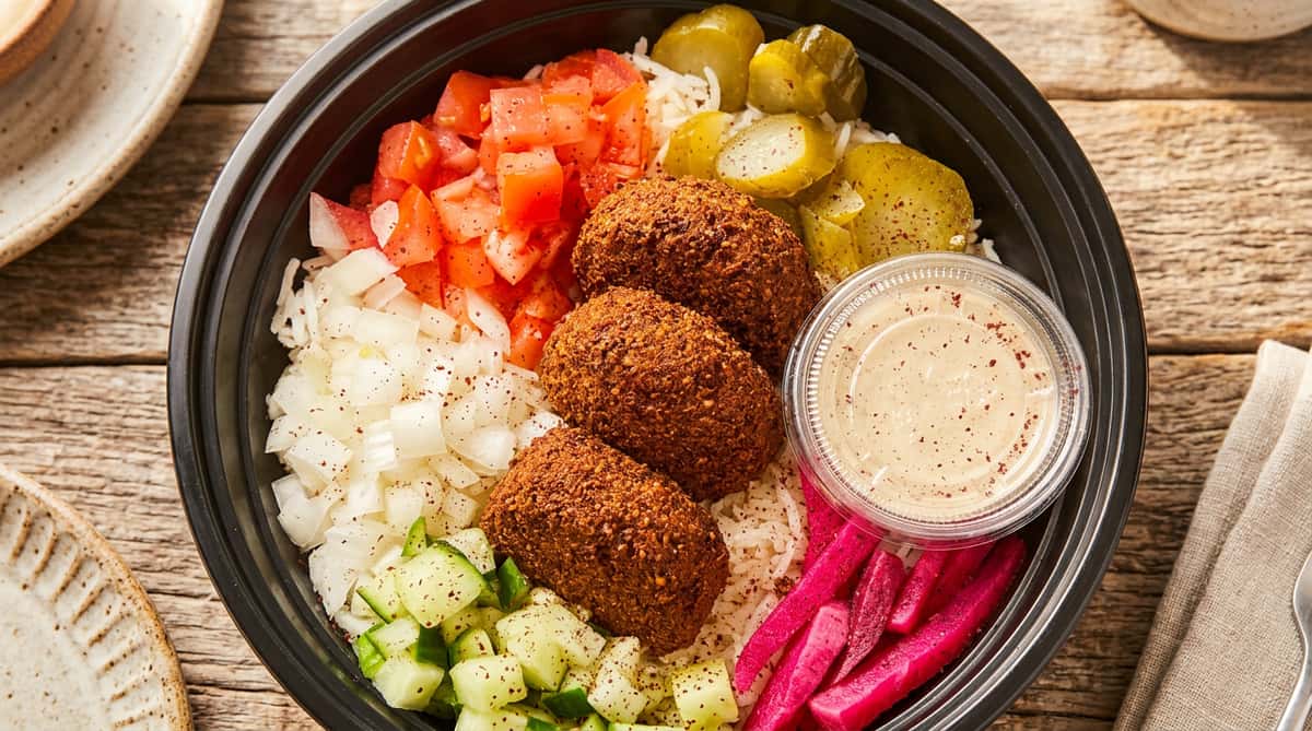 Falafel bowl with rice, diced tomatoes, onions, cucumbers, pickles, pickled turnips, and a side of sauce.