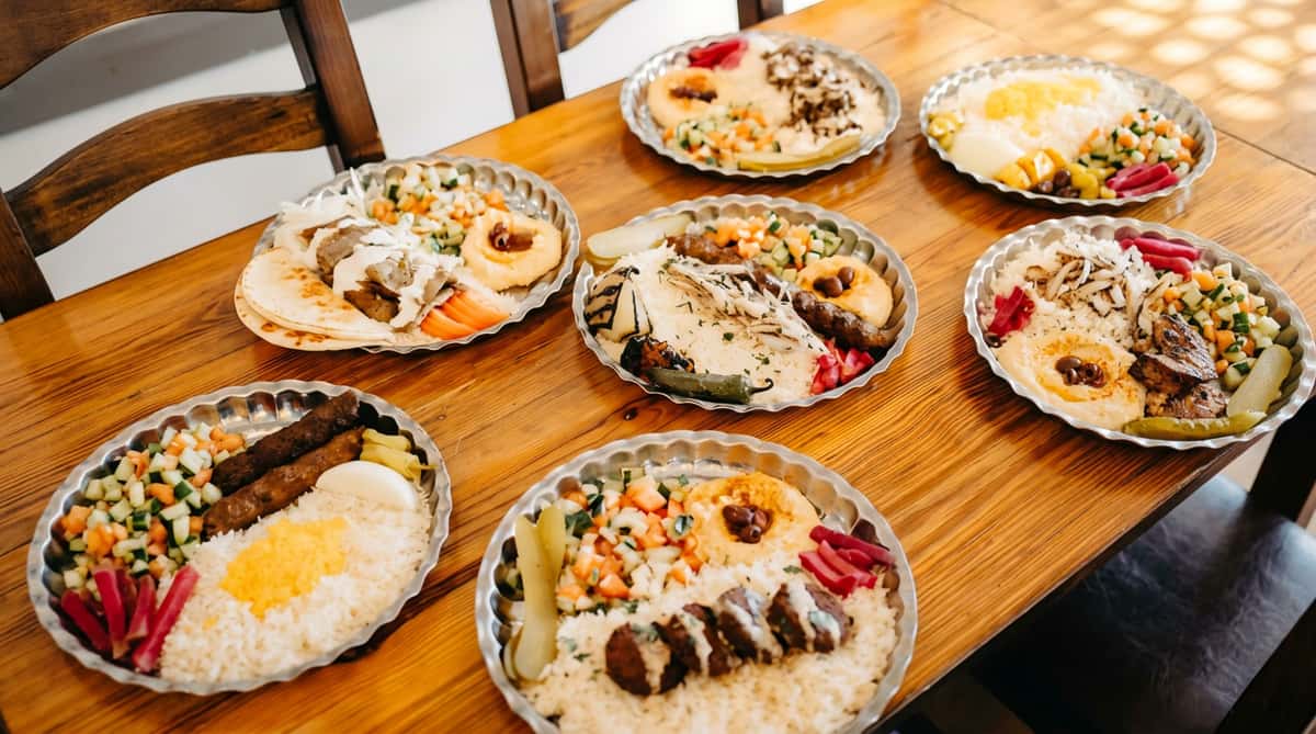 Assortment of Middle Eastern platters with rice, hummus, meats, falafel, salads, and pickles on a wooden table.