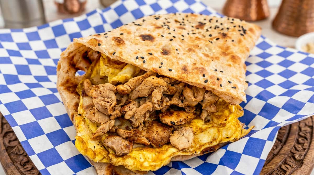 Flatbread sandwich filled with seasoned meat, scrambled egg, and cabbage, served on blue and white checkered paper.