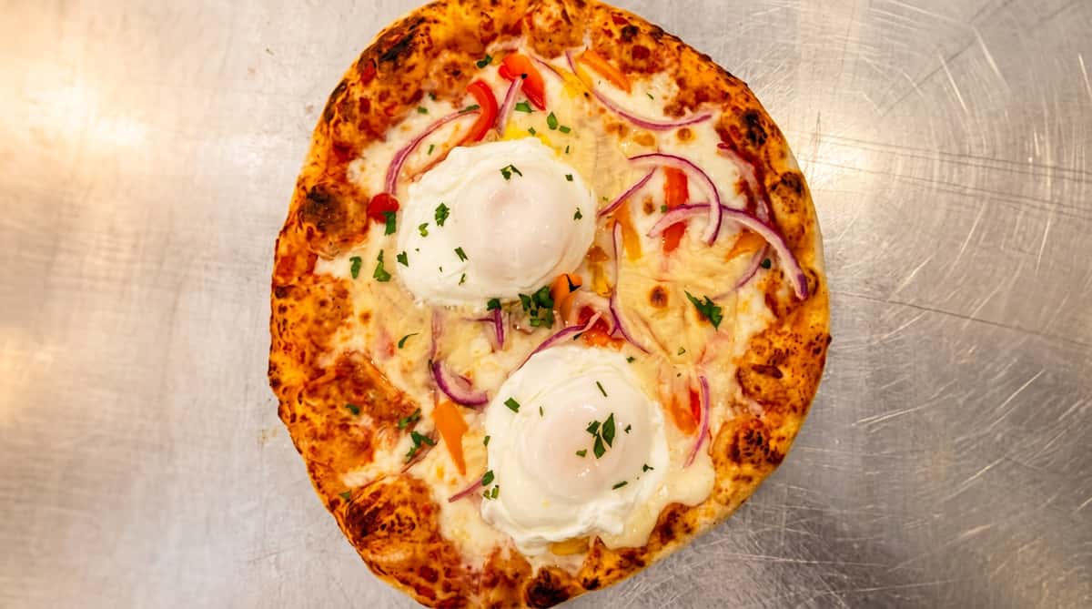 Pizza topped with two poached eggs, melted cheese, red onion, colorful bell peppers, and fresh herbs.