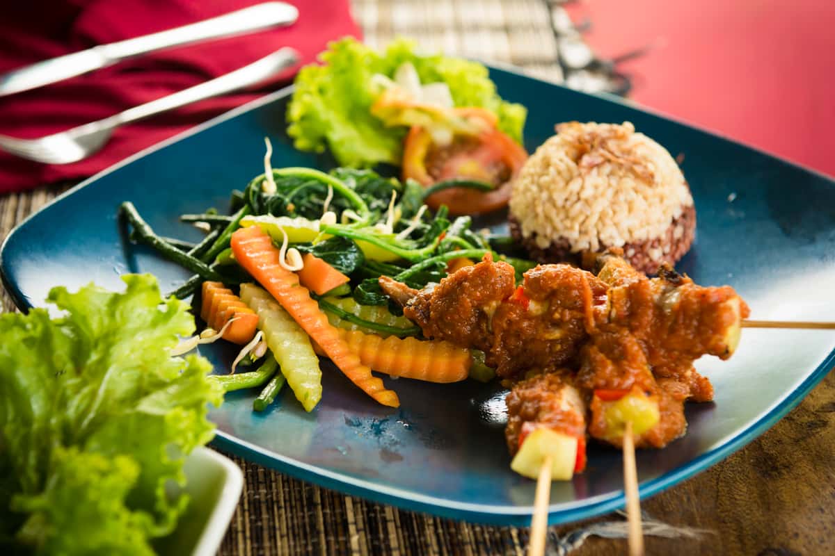 A vibrant plate of Indonesian-inspired skewers (Satay) with rice and fresh vegetables, representing the international influences and healthy options on the New American/European fusion menu.