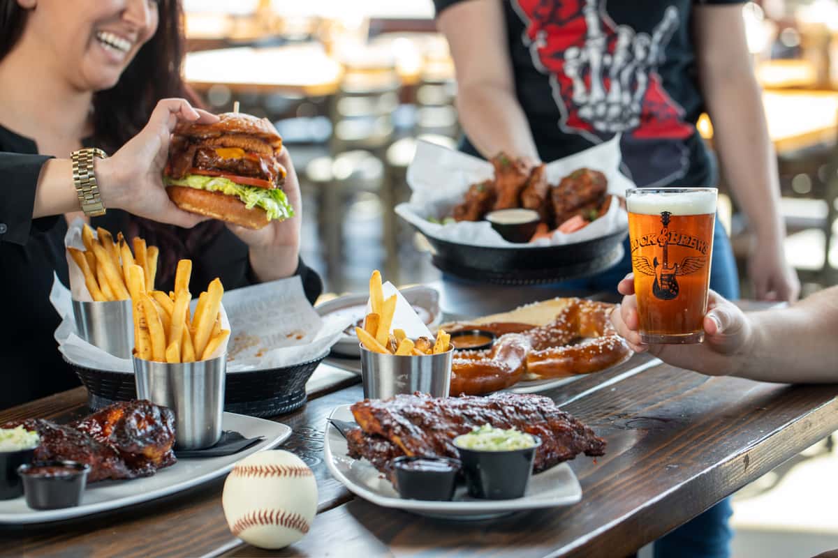 Guests enjoying burgers, ribs, fries, pretzels, wings, and beers at Rock & Brews, with a baseball on the table and large "BASEBALL" text over the image.
