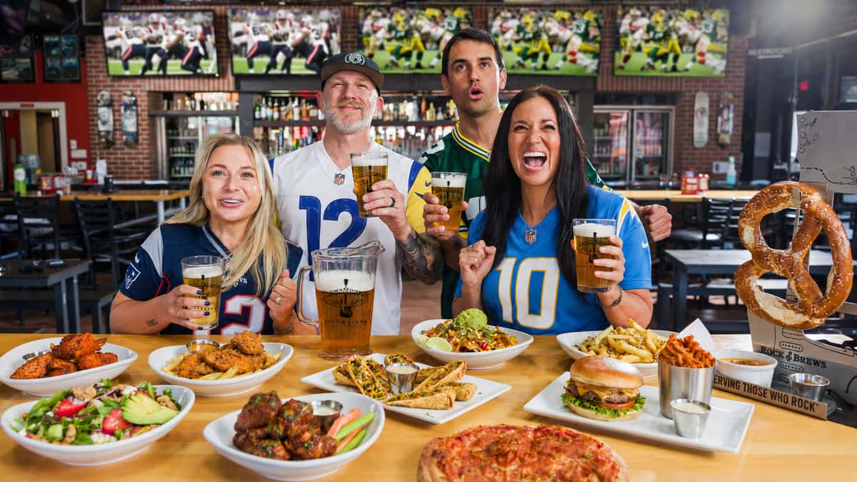 Football fans in Patriots, Rams, Chargers, and Packers jerseys cheering at Rock & Brews. The group has beers in their hand and a full spread of food on the table in front of them while football plays on the screens around them.