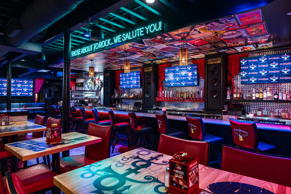 Rock & Brews first-floor dining room with red booths, branded bar seating, beer taps, TV screens, colorful rock posters on the ceiling, and a teal neon sign that reads "For those about to rock, we salute you!"