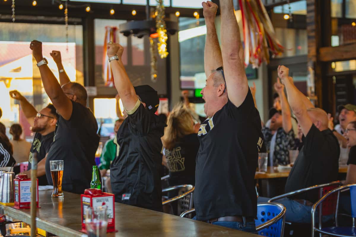 Guests cheering with their arms raised at a Rock & Brews bar, with drinks on the counter and large "MORE" text over the image.