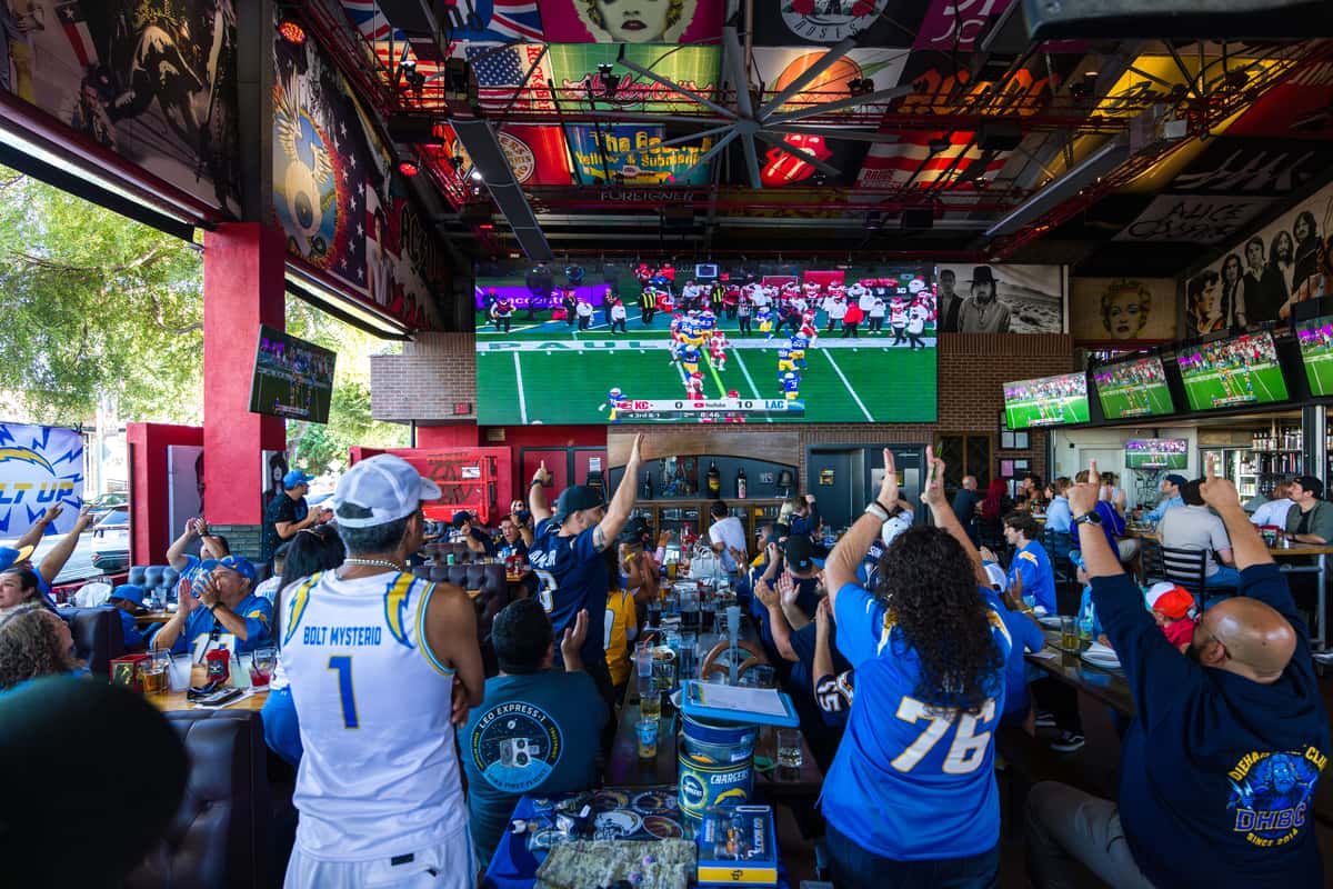 Chargers fans cheering inside Rock & Brews El Segundo while watching a live NFL game on a massive 28-foot LED wall, surrounded by rock-and-roll artwork and a high-energy sports bar atmosphere.
