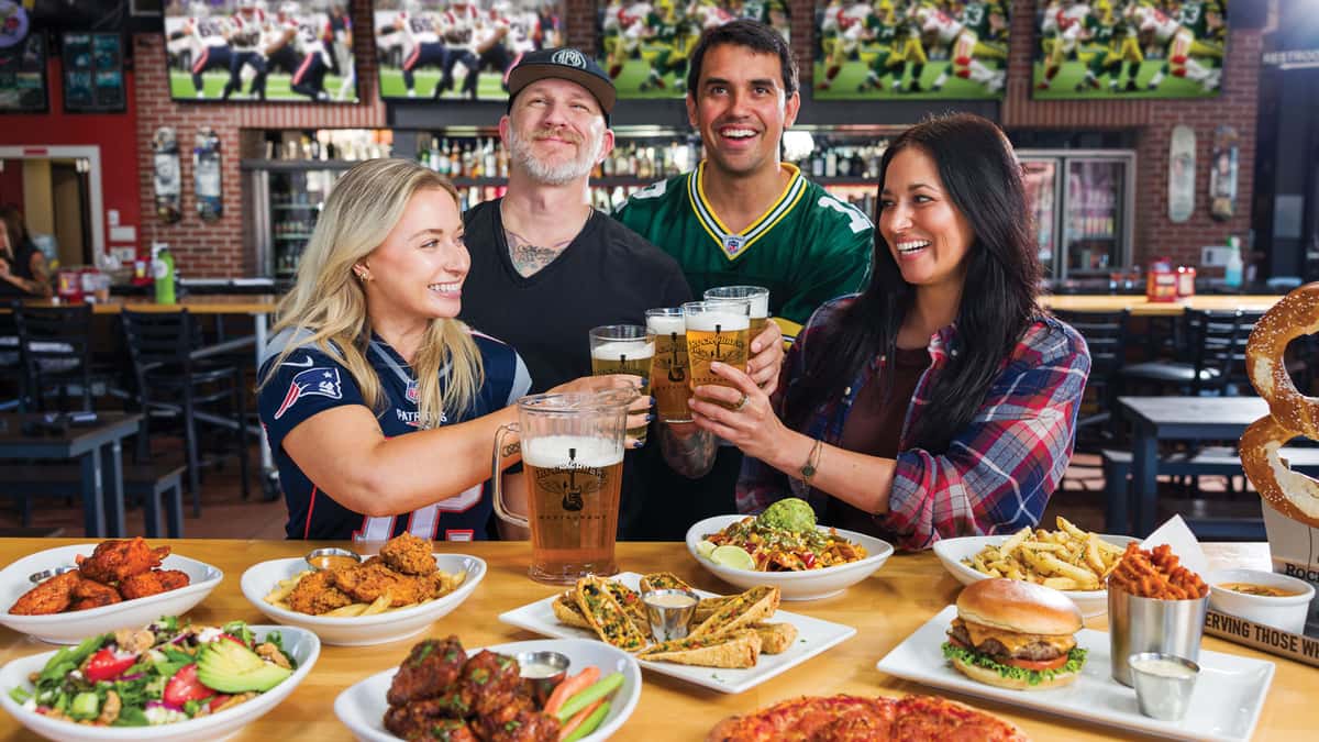 Four guests cheering beers at a Rock & Brews table filled with wings, salads, burgers, fries, pizza, and appetizers, with football games playing on TVs in the background.