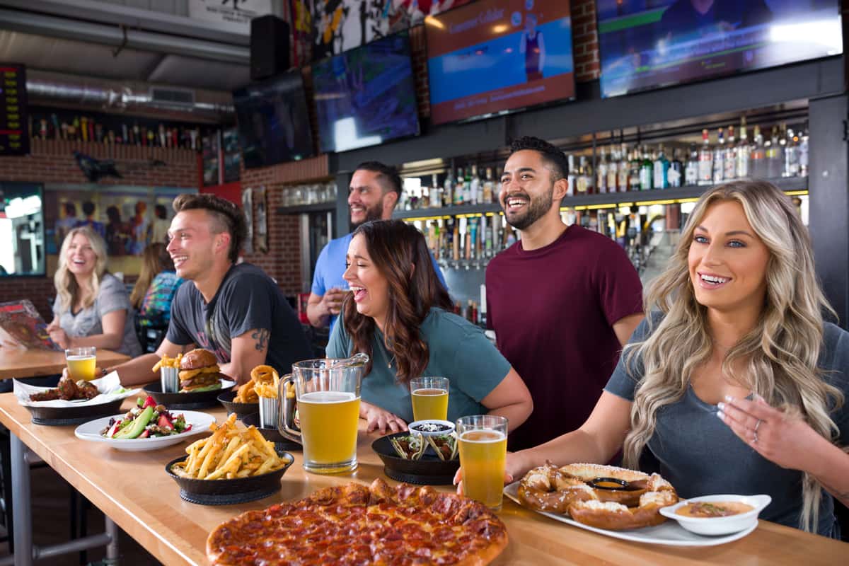 A group of friends laughing and watching TVs while seated at a long bar. The counter is filled with a variety of pub food, including a pepperoni pizza, a large soft pretzel, fries, tacos, and burgers, alongside several glasses and a pitcher of beer. The setting is a vibrant sports bar with a friendly, social atmosphere.