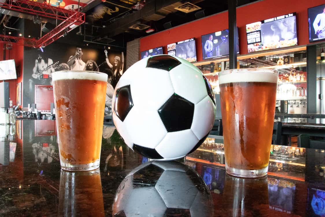 Soccer ball and two beers on a Rock & Brews bar, with TVs and rock-and-roll decor in the background and large "SOCCER" text over the image.