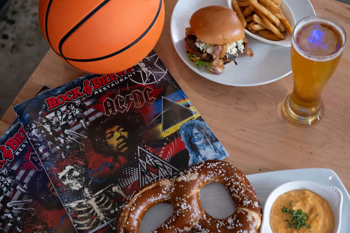 Basketball-themed Rock & Brews table with a basketball, burger, fries, beer, giant pretzel, and rock-and-roll menu, with large "BASKETBALL" text over the image.