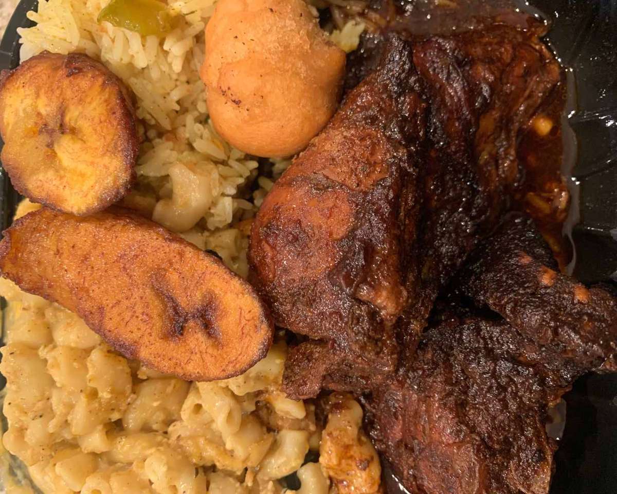 Jamaican meal with jerk chicken, fried plantains, rice, mac and cheese, and a festival dumpling on a black plate.