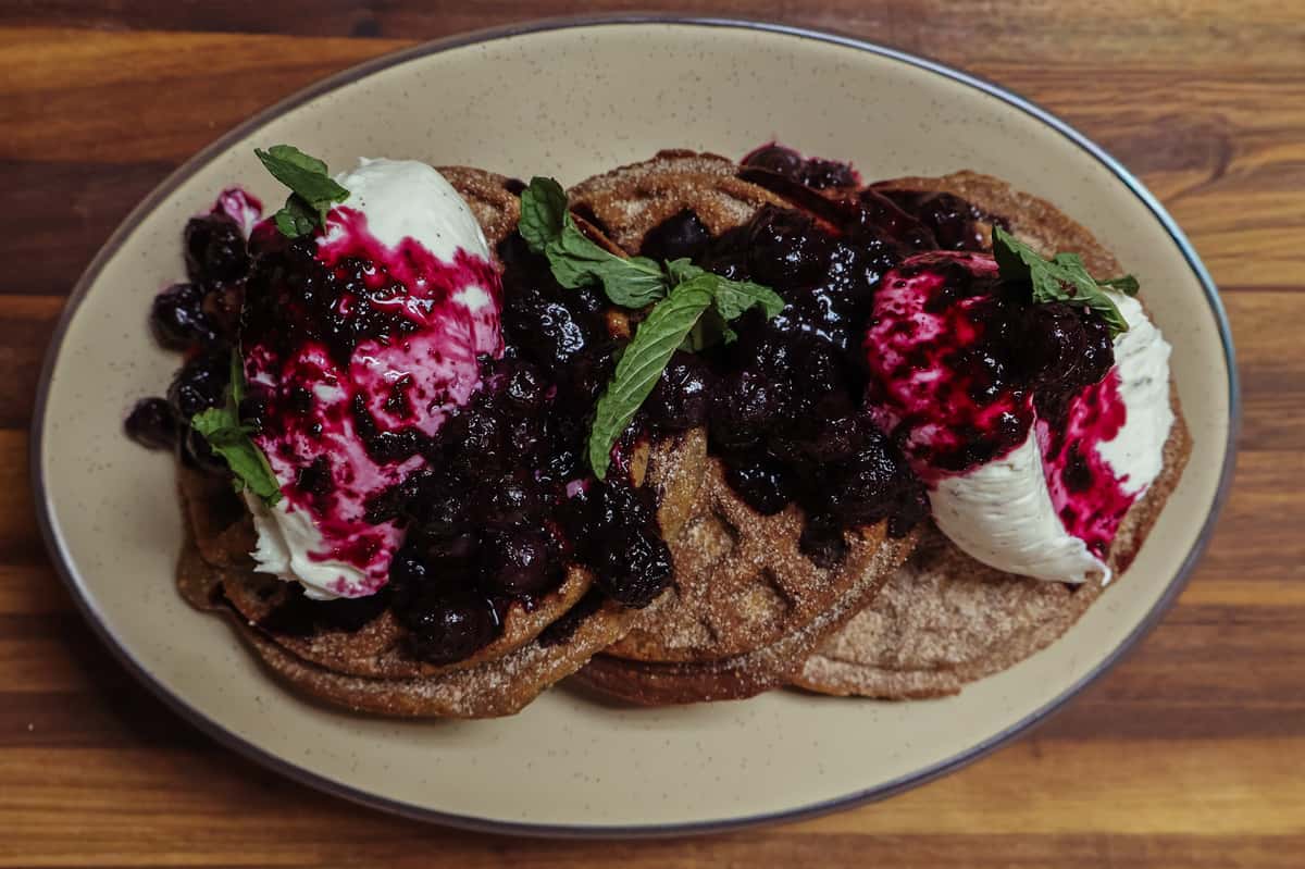 This image showcases a delicious dessert dish featuring crispy waffles dusted with cinnamon sugar, generously topped with vanilla ice cream or whipped cream, a vibrant berry compote (likely blueberry), and fresh mint leaves for garnish. The contrast between warm waffles and cold cream, plus the sweet and tart berries, makes it visually and flavorfully indulgent. Write alt text descriptions for the image(s) attached. If keywords are provided include them naturally. Do not use terms such as "image of", be descriptive and specific. If the focus of the image is unclear, provide a general description. The alt text description should be 125 characters or less. Output only the image description. Grilled chicken bowl with sliced avocado, black beans, rice, sautéed peppers and onions, queso fresco, and fresh cilantro on a wooden table. Baked casserole topped with melted cheese, fresh cilantro, and crumbled queso fresco in a metal skillet on a wooden surface. Grilled breakfast quesadilla filled with eggs, cheese, and meat, served with salsa verde and sour cream on a ceramic plate. Cheesy baked casserole topped with cilantro and queso fresco beside blue corn enchiladas filled with vegetables in metal skillets. Grilled steak tacos with sautéed onions and cheese, served with rice, roasted vegetables, fresh cilantro, and creamy dipping sauce. Two blue corn tacos topped with shredded lettuce, onions, and diced pickles, served with rice, black beans, and a side of red salsa. Three corn husk-wrapped tamales served with green tomatillo salsa and red chili sauce on a ceramic plate. Sliced grilled steak served over rice with avocado, sautéed peppers, black beans, greens, and zucchini ribbons in a bowl. Braised beef short ribs served over creamy grits with sautéed peppers, tomatoes, and fresh cilantro garnish.