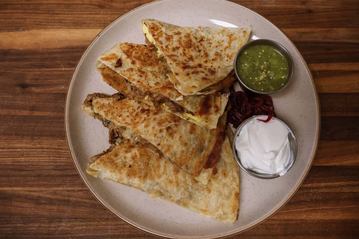 Grilled breakfast quesadilla filled with eggs, cheese, and meat, served with salsa verde and sour cream on a ceramic plate.