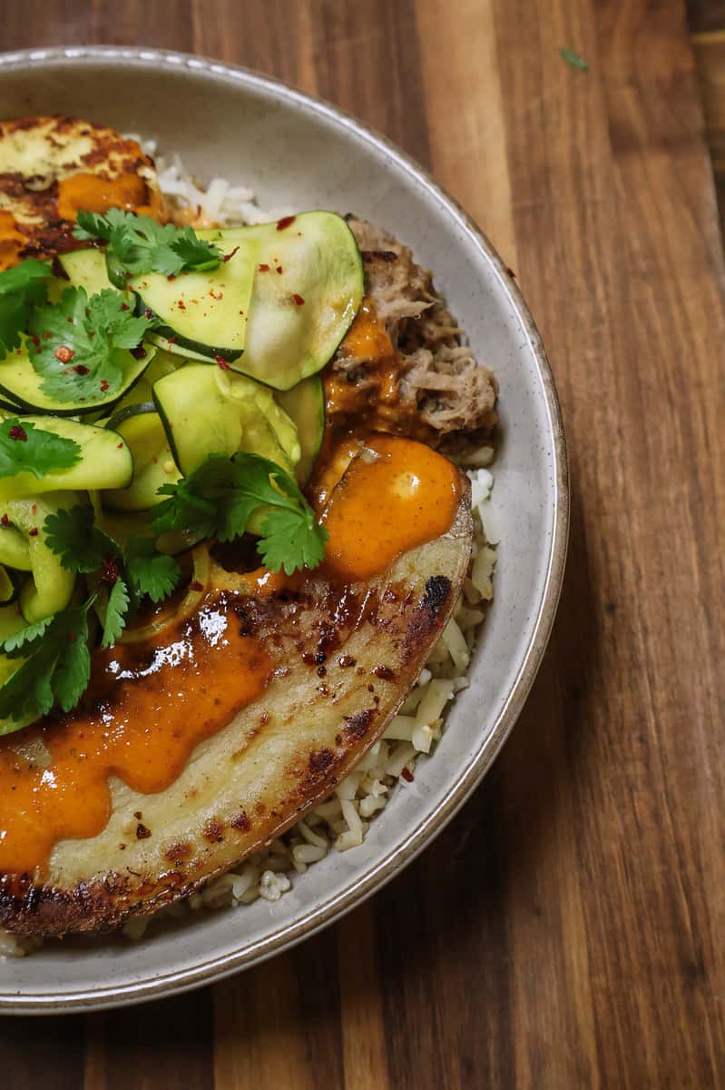 Pork and rice bowl with fried plantain, squash ribbons, cilantro, and spicy orange sauce, served with a green dipping sauce.