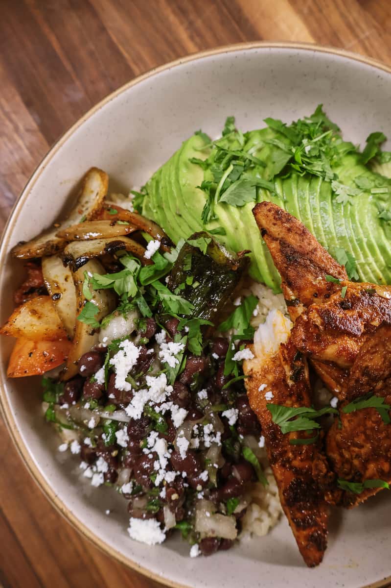 Grilled chicken bowl with sliced avocado, black beans, rice, sautéed peppers and onions, queso fresco, and fresh cilantro on a wooden table.