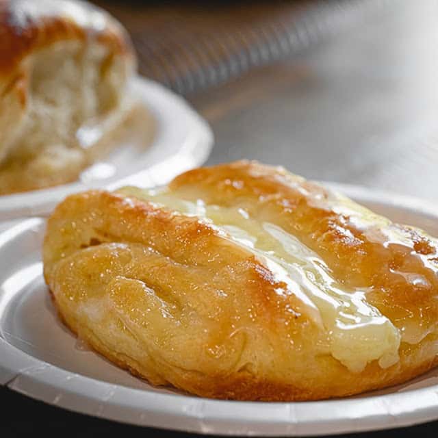 Glazed pastry with golden layers and sweet icing drizzle served on a white paper plate, offering a flaky and indulgent breakfast treat.