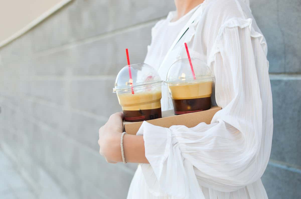 Woman in a white blouse holding a coffee tray with two iced espressos topped with thick foam, captured outside a trendy café.