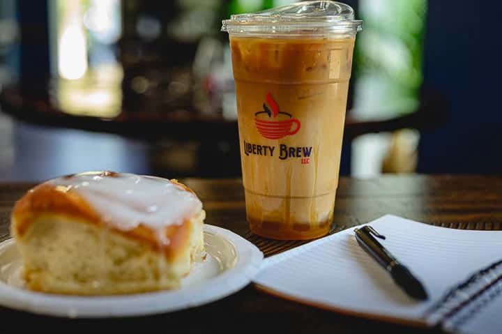 Iced coffee and a soft cinnamon roll with icing on a paper plate, next to a notepad and pen at Liberty Brew—perfect for a cozy café work session.