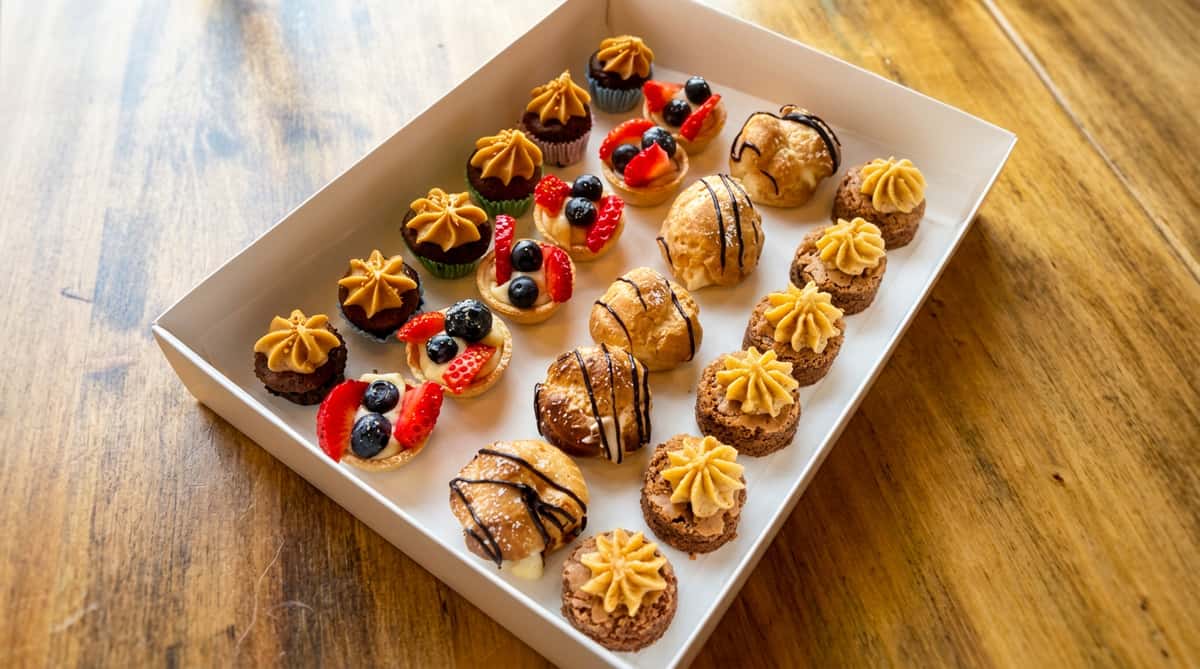 Assorted mini desserts in a white box, including fruit tarts, cream puffs, and brownies with piped frosting.