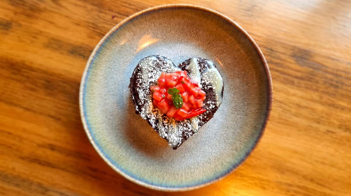 Heart-shaped chocolate dessert with white drizzle, powdered sugar, diced strawberries, and a mint sprig on a gray plate.