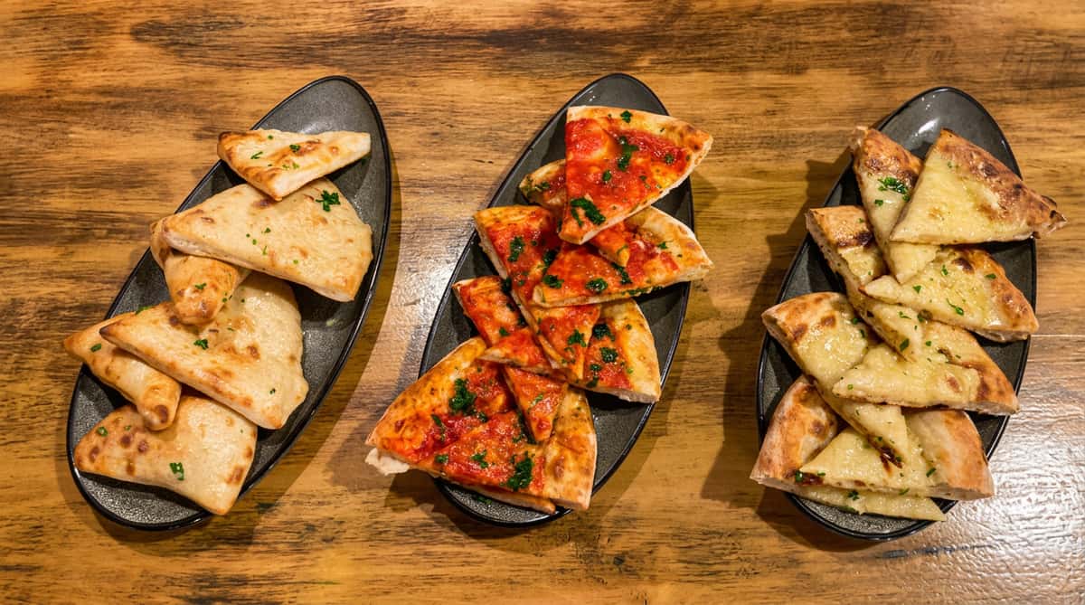 Assorted flatbreads: plain, with tomato sauce and herbs, and with melted cheese and herbs, on a wooden table.