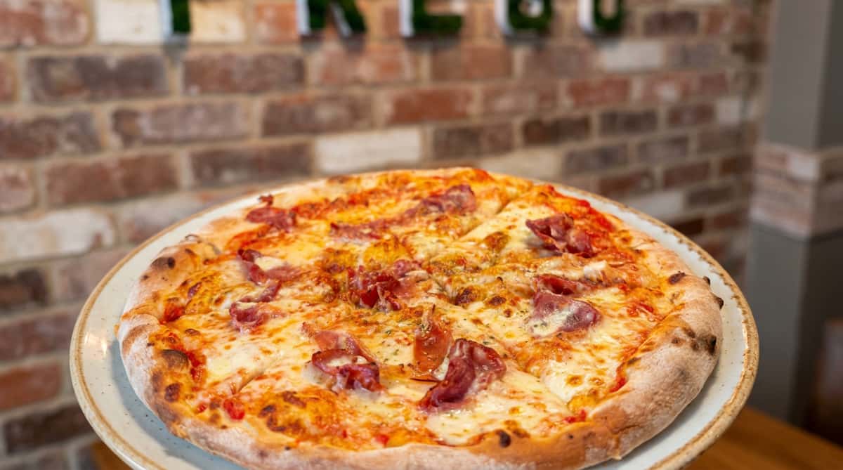 Salami pizza with melted cheese and red sauce on a white plate, with a brick wall and "PREGO" sign in the background.