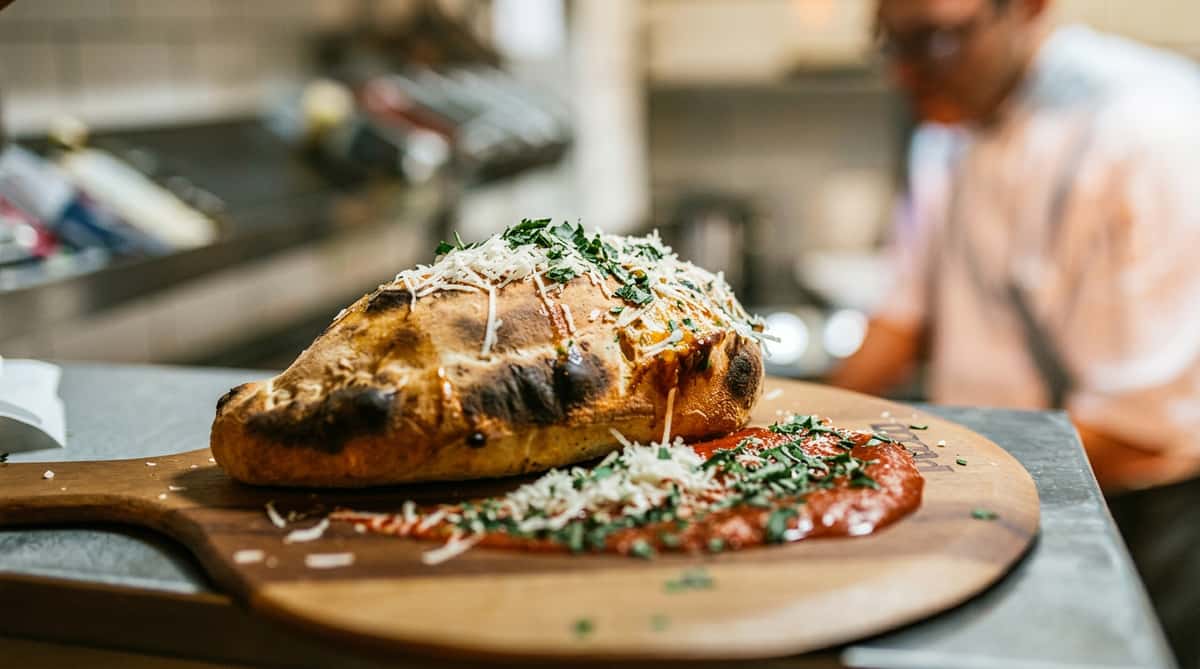 Calzone with melted cheese and green herbs, marinara sauce on a wooden pizza peel.