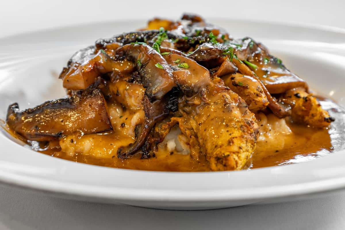 Blackened chicken breast topped with sautéed mushrooms and smothered in a rich, savory brown gravy, served over creamy mashed potatoes on a white plate.