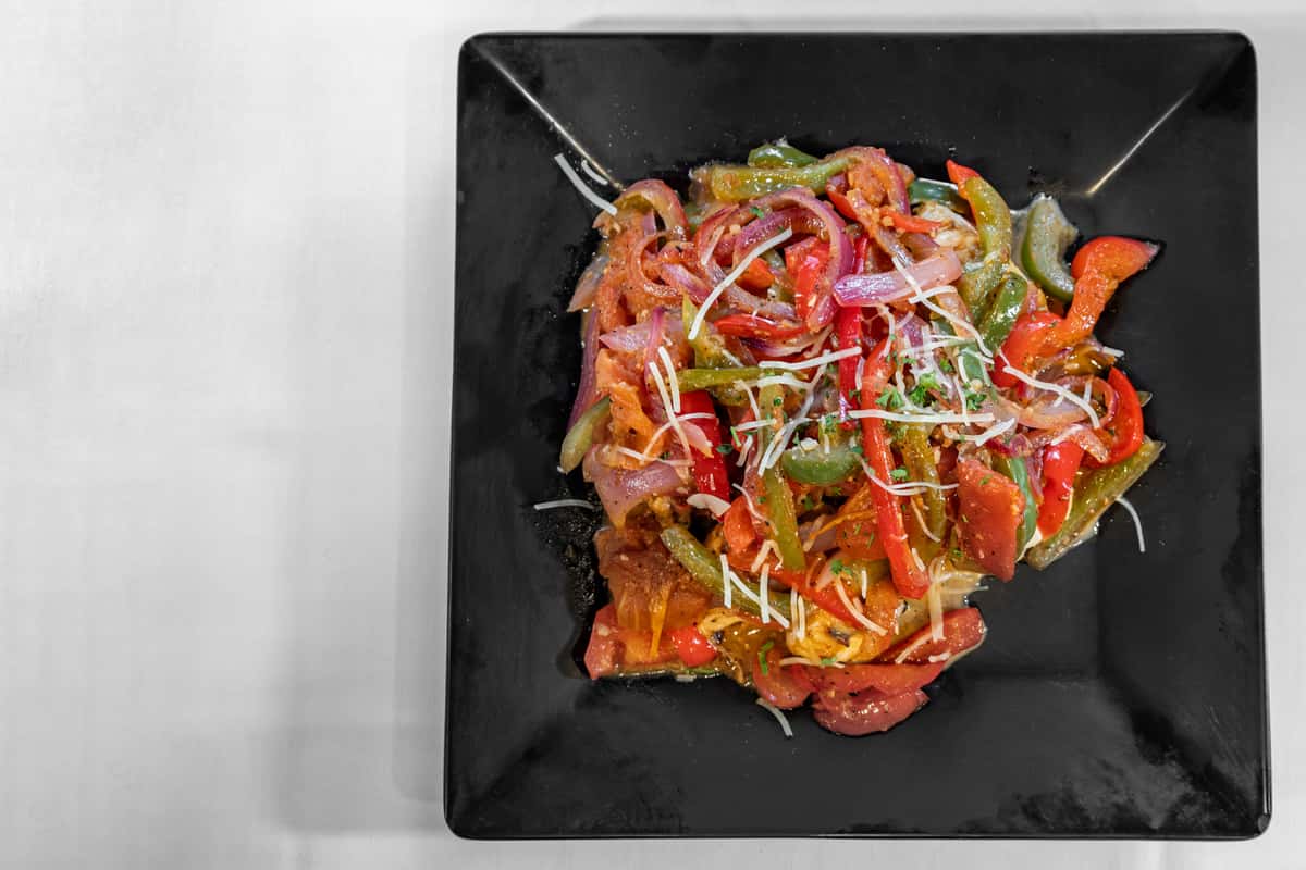 Colorful sautéed peppers and onions served over a grilled protein base, topped with shredded parmesan and fresh herbs, plated on a sleek black dish for a vibrant and flavorful entrée.