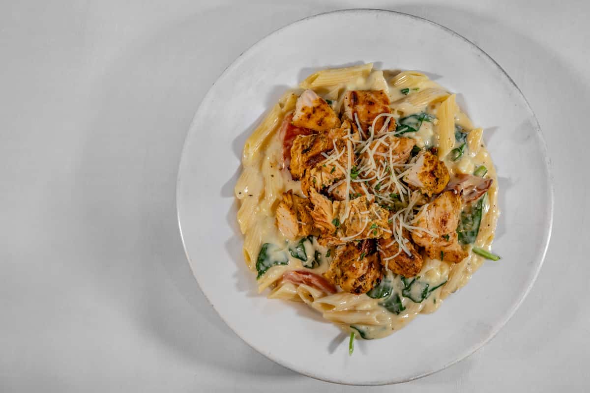 Close-up of creamy chicken Alfredo pasta with grilled chicken, penne noodles, sautéed spinach, and shredded parmesan cheese, served on a white plate in a casual dining setting.