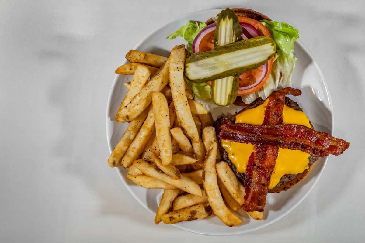 Classic bacon cheeseburger topped with melted cheddar, crispy bacon, lettuce, tomato, red onion, and pickles, served with seasoned French fries on a white plate.