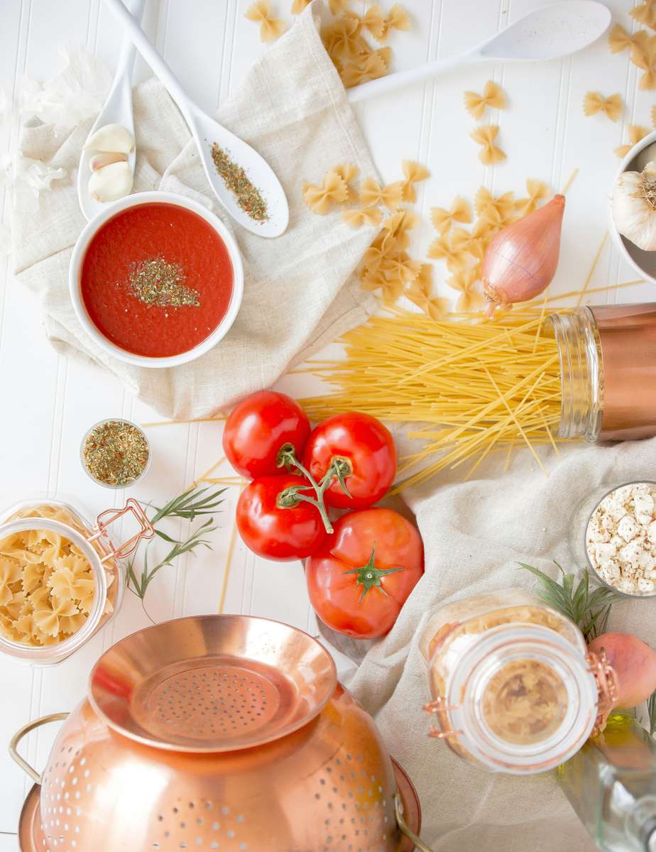 Flat lay of classic Italian pasta ingredients including dry spaghetti, farfalle, vine tomatoes, tomato sauce, herbs, garlic, shallot, and a copper colander on a white wooden surface.