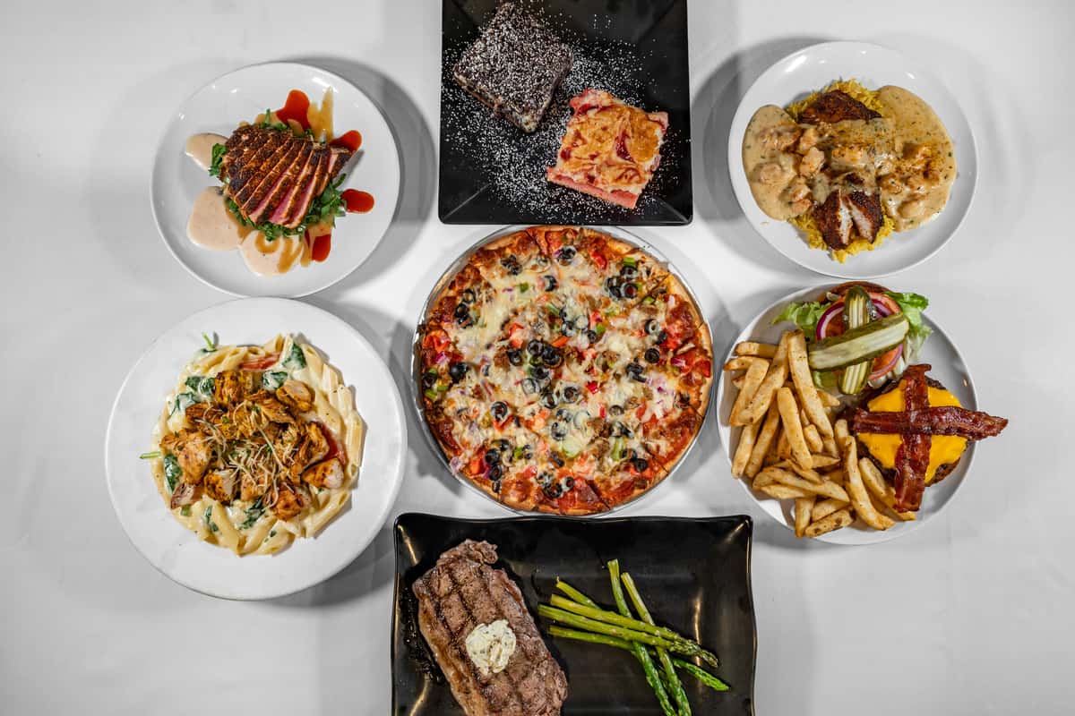 Overhead view of a diverse American restaurant spread featuring a fully loaded veggie pizza, grilled ribeye steak with asparagus, creamy chicken Alfredo pasta, bacon cheeseburger with fries and pickles, seared ahi tuna over greens with sauce drizzle, blackened fish topped with shrimp in creamy sauce over rice, and a dessert plate with a chocolate brownie and fruit-topped pastry.