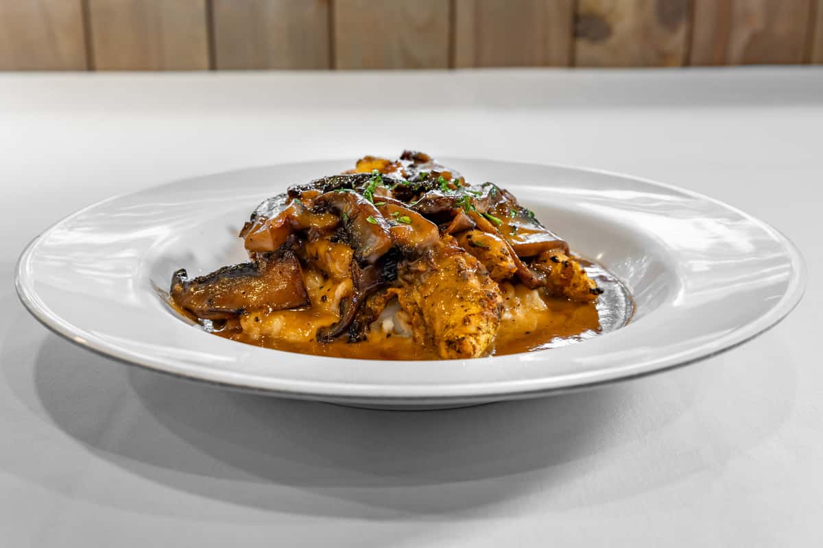 Grilled blackened chicken breast topped with sautéed mushrooms and rich brown gravy, served over creamy mashed potatoes on a white dinner plate.