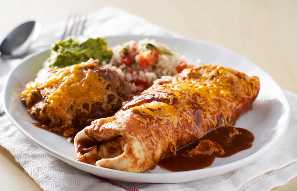 A plated meal featuring two enchiladas topped with melted cheese and a side of rice and guacamole.