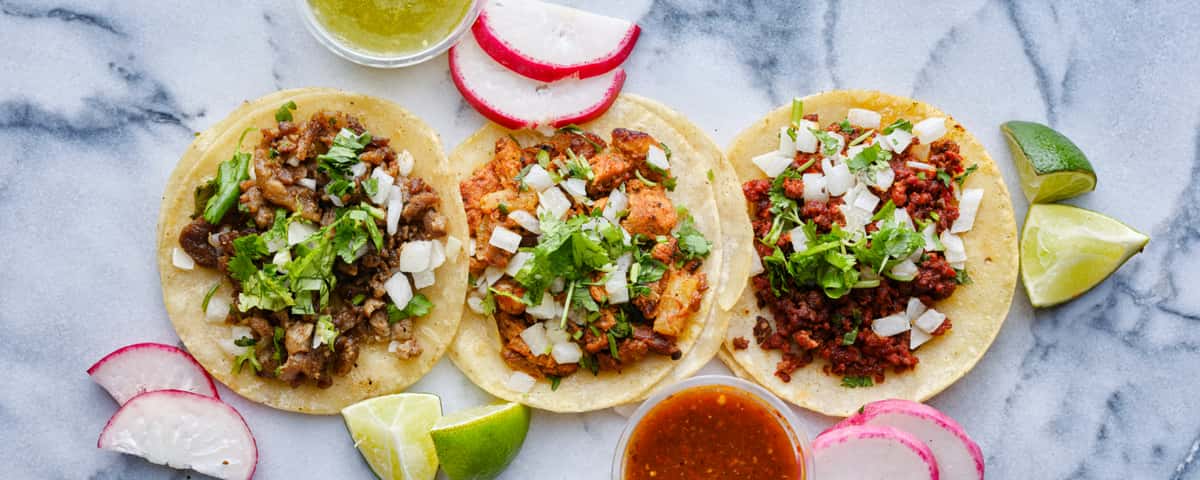 An assortment of three street tacos on a white marble table with salsa and toppings
