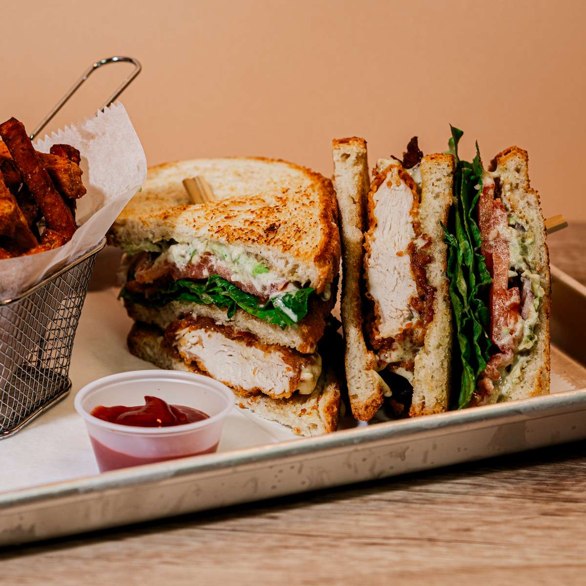 A hearty triple-decker chicken club sandwich featuring grilled chicken, crisp bacon, lettuce, and tomato, sliced and served with a side of golden french fries and dipping sauce.