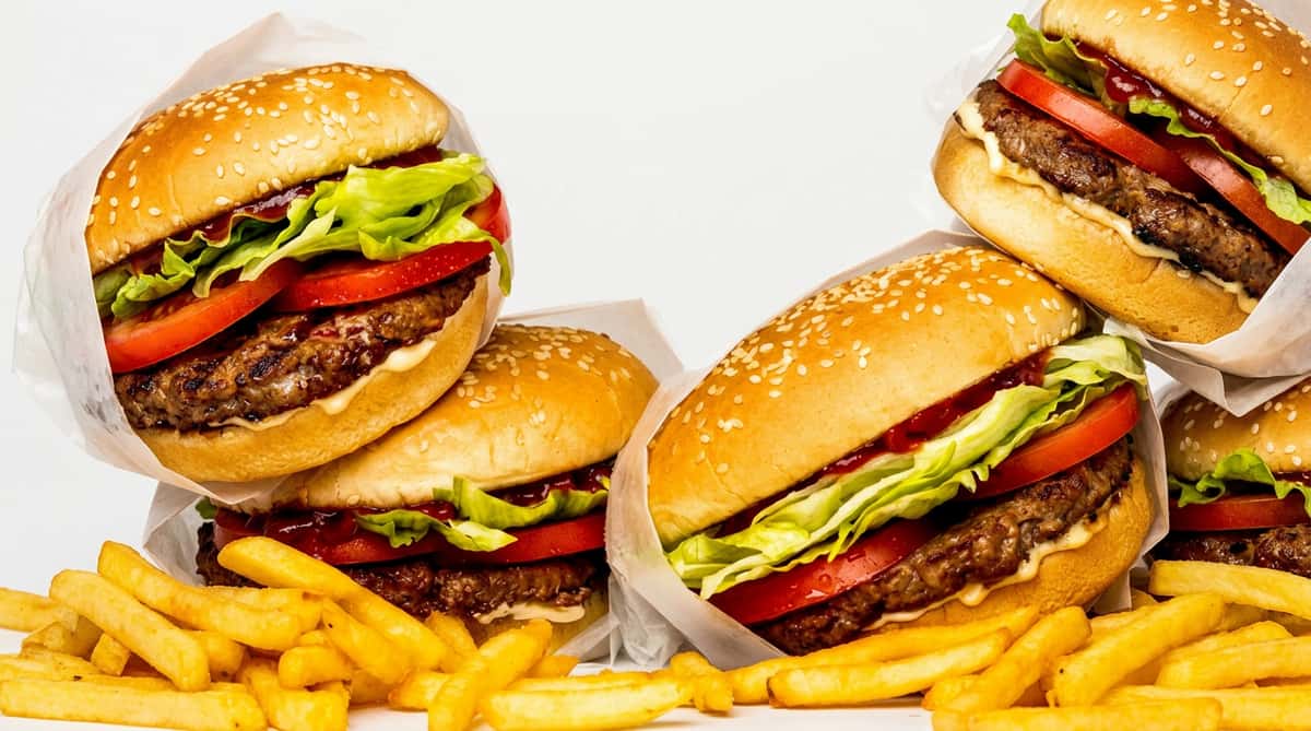Three sesame seed burgers with beef patties, lettuce, tomato, and white paper wrapping, alongside golden french fries.