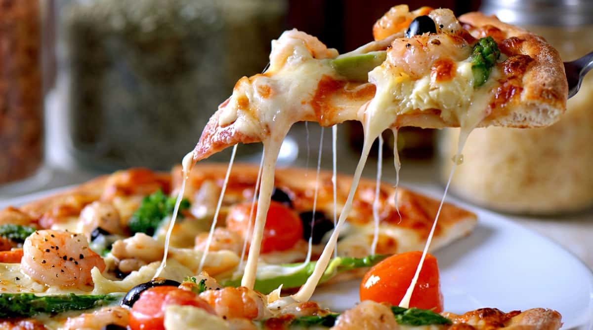 Pizza slice with melted cheese stretching, topped with shrimp, cherry tomatoes, and black olives.