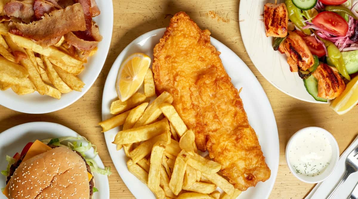 Fried fish and chips with lemon, alongside plates of kebab meat and grilled chicken salad.