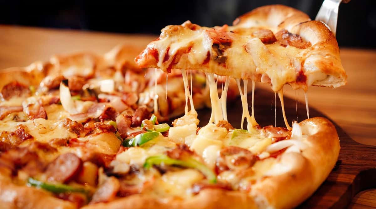 Pizza slice lifted from a whole pizza, with melted cheese stretching, meat, onions, and green peppers visible.