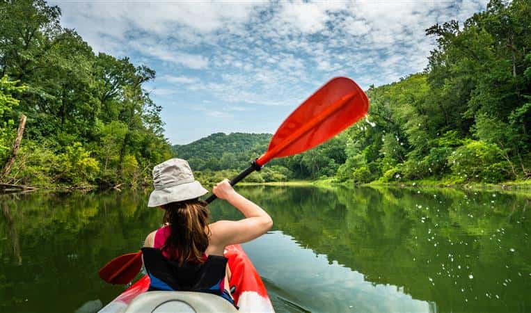 girl paddling down a river on a kayak with trees around her and a blue sky filled with puffy clouds on a sunny day