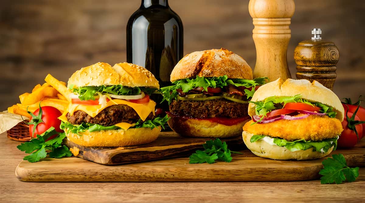 Three burgers: a cheeseburger, a beef burger with bacon and pickles, and a chicken burger, with fries and tomatoes.