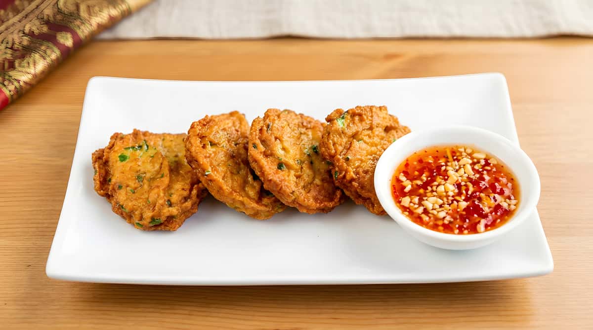 Four fried fish cakes with a side of sweet chili dipping sauce on a white rectangular plate.