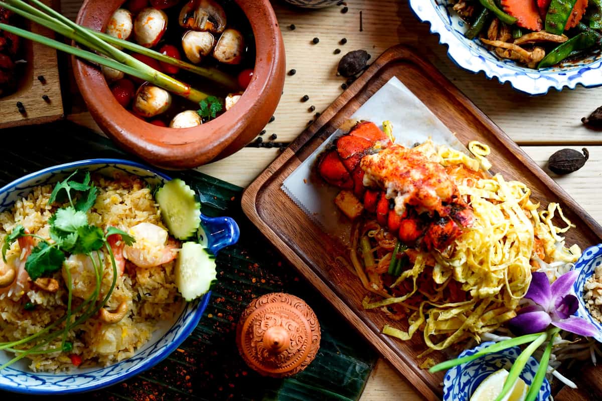 Assorted Asian dishes including lobster noodles, shrimp fried rice, and spicy soup with mushrooms and herbs on a rustic table