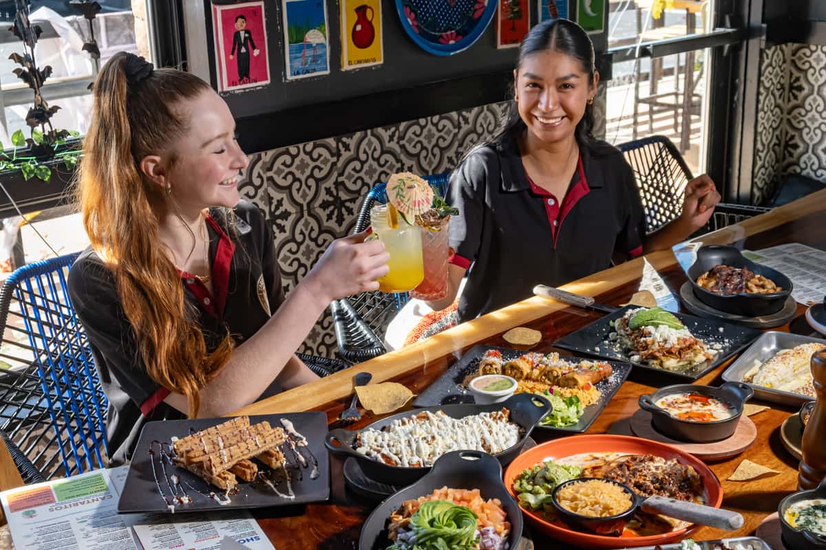 Two women smiling and toasting colorful cocktails over a table filled with vibrant Mexican dishes in a festive restaurant setting.