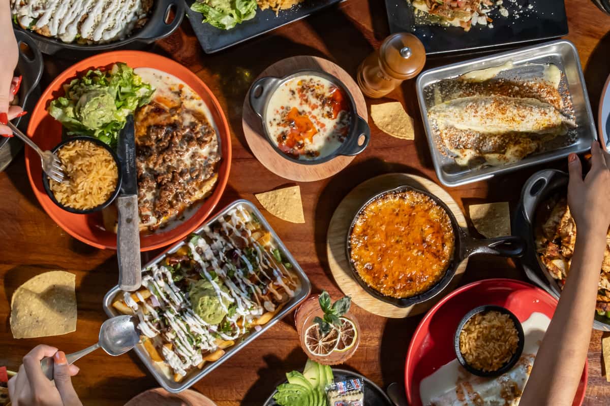 Assorted Mexican dishes including enchiladas, queso dip, and guacamole fries served on a wooden table with tortilla chips.