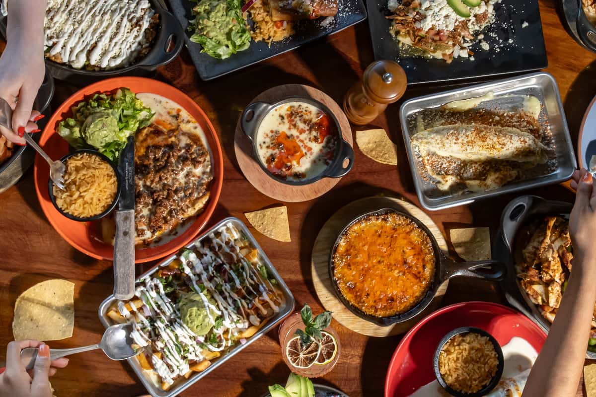 Assorted Mexican dishes including enchiladas, queso dip, and guacamole fries served on a wooden table with tortilla chips.