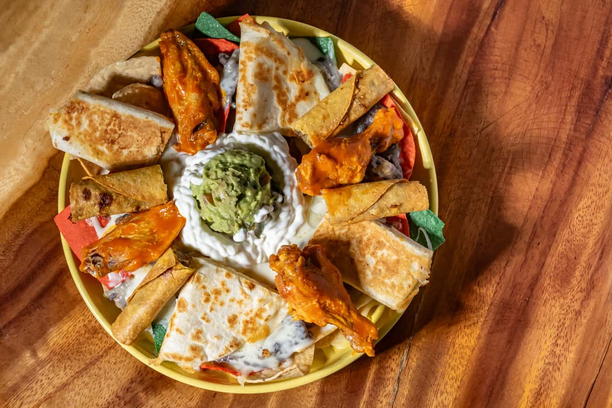 This dish is a festive appetizer platter featuring a mix of Mexican favorites — crispy taquitos, cheesy quesadilla wedges, and saucy buffalo wings — all arranged around a center of creamy guacamole and sour cream, served over colorful tortilla chips for a vibrant presentation.